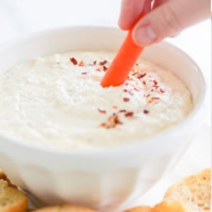 A hand dips a carrot stick into a luxurious Asiago Cheese Dip, garnished with red pepper flakes and surrounded by slices of bread.