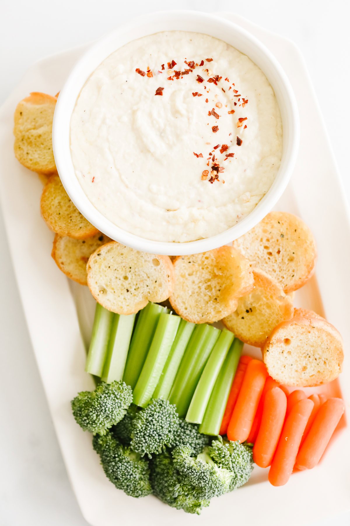 A bowl of creamy Asiago Cheese Dip garnished with red flakes, surrounded by sliced bread, broccoli, celery, and carrots on a rectangular white plate.