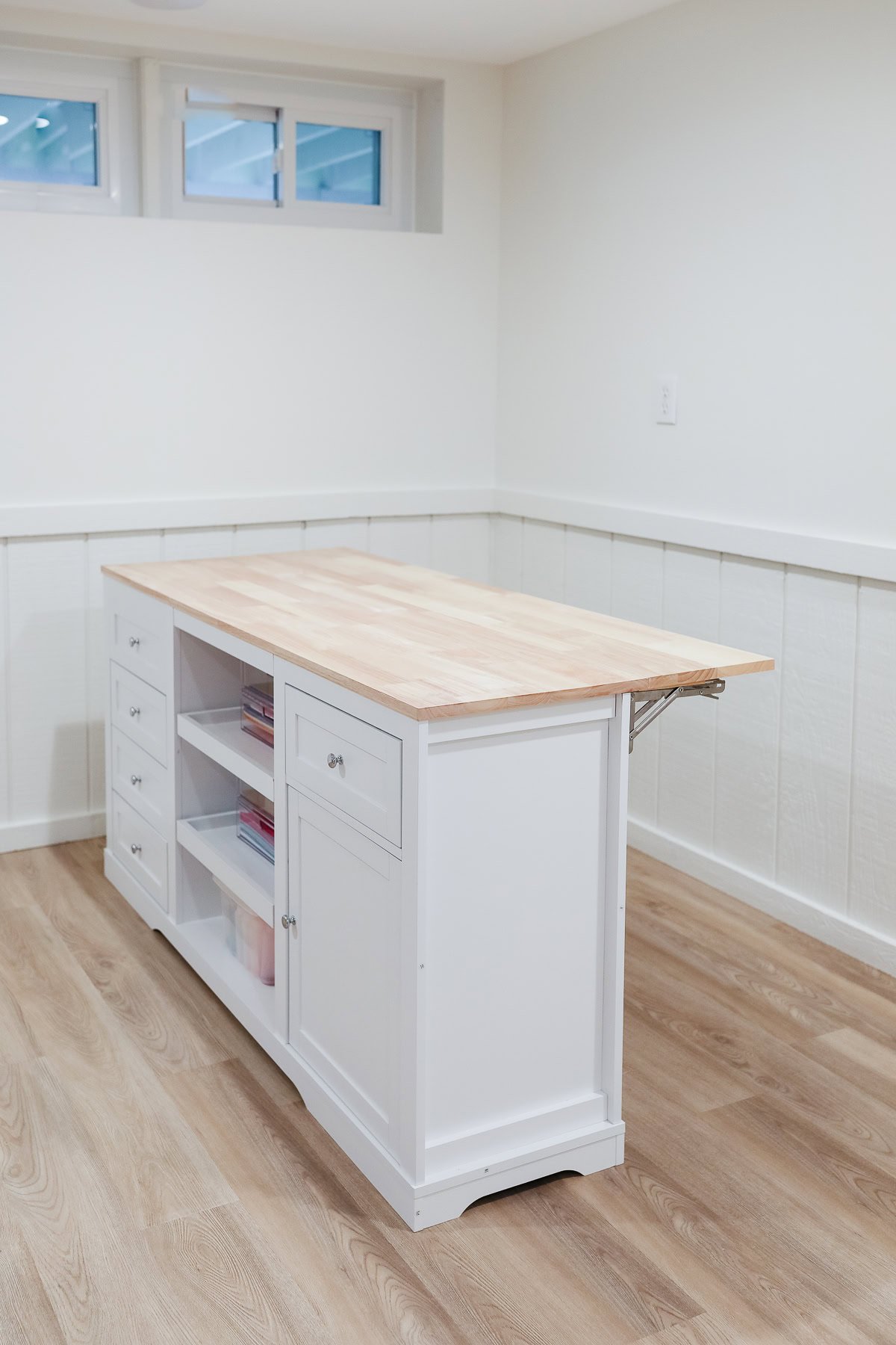 A white kitchen island with a wooden top, perfect for craft space ideas, features open shelves and drawers. It's set against a white wainscoted wall and light wood floor in a bright room.