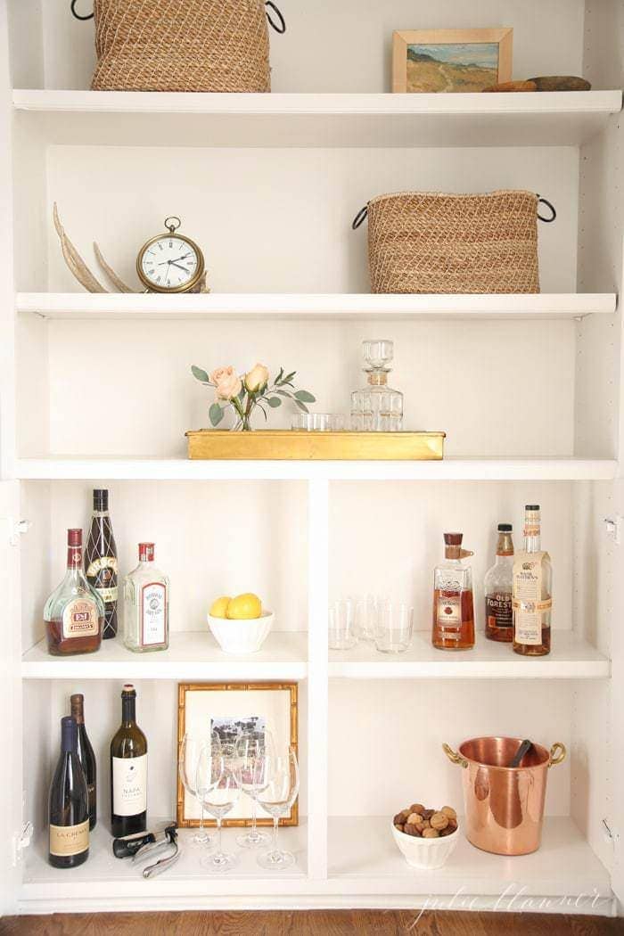 A white shelf artfully combines organizing crafts with decorative baskets, a clock, bottles of alcohol, wine glasses, lemons, and a copper ice bucket.