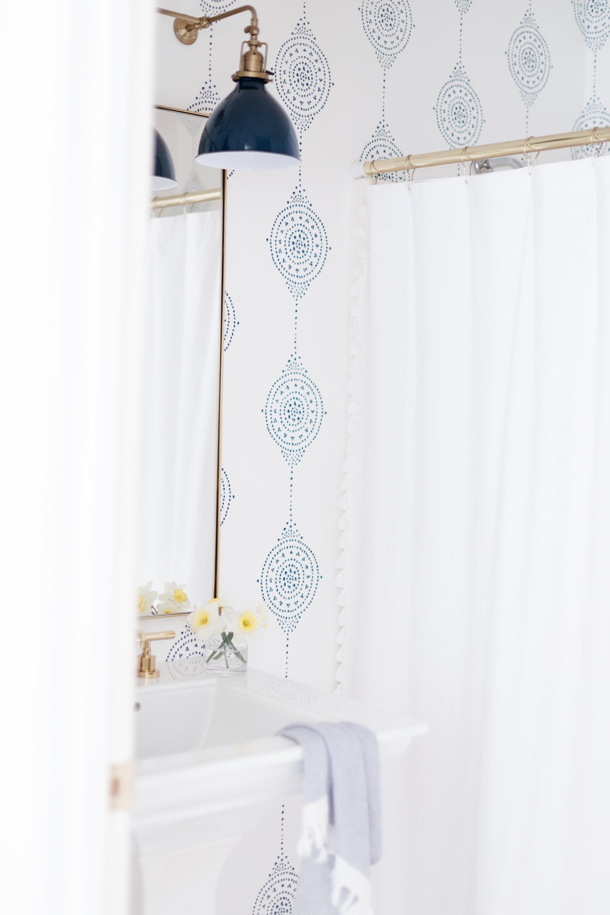 The bathroom features a Serena and Lily wallpaper, complementing the white shower curtain, while a dark blue light fixture casts a cozy glow. The sink is elegantly adorned with a soft towel and fresh flowers.