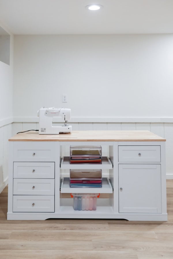 A sewing machine sits on a wooden and white storage table with drawers and shelves, showcasing craft space ideas. It neatly holds various colored paper and a plastic container in a well-lit room with wooden flooring.