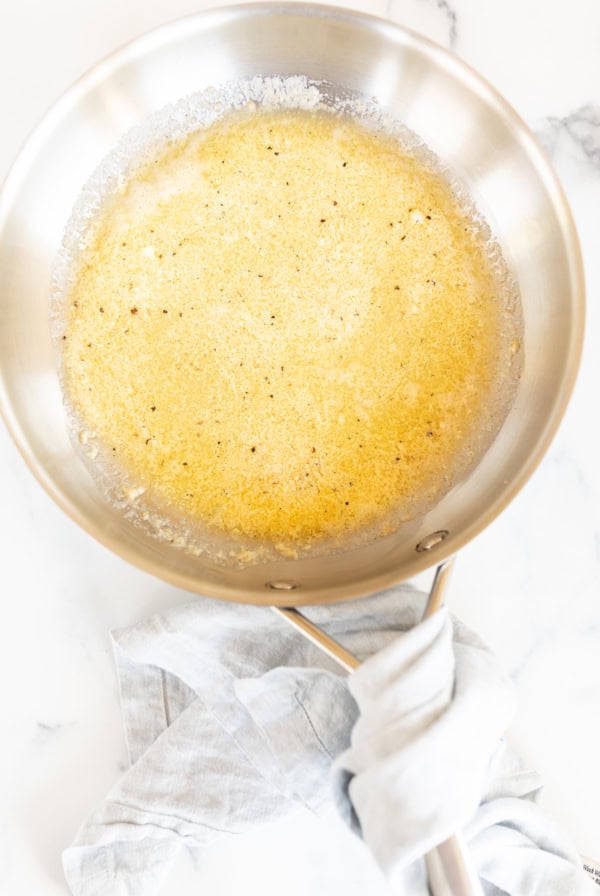 Stainless steel pan with melted butter bubbling, a hint of garlic parmesan sauce in the air, sits on a marble surface with a blue kitchen towel wrapped around the handle.
