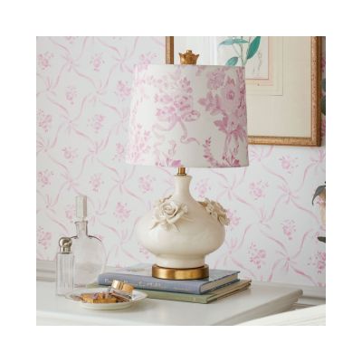 A decorative table lamp with a floral pink and white shade sits on books next to a glass bottle and a small dish with cookies. Channeling a LoveShackFancy look for less, the scene is set against a backdrop of charming floral-patterned wallpaper.