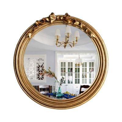 A LoveShackFancy look for less comes to life as a round gold-framed mirror elegantly reflects a dining room featuring a chandelier, a vase on the table, and white cabinets in the background.