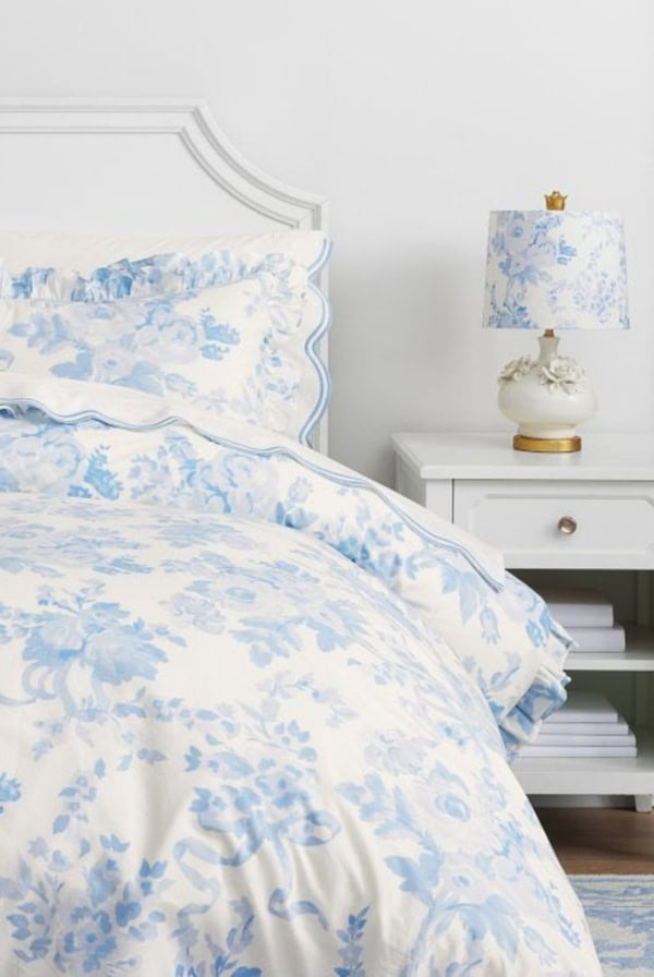 The bedroom scene exudes a LoveShackFancy look for less, showcasing a white bed adorned with blue floral bedding. A matching lamp graces the white nightstand, adding charm and cohesion to the stylish yet budget-friendly aesthetic.