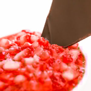 Close-up of diced red strawberries with a piece of dark chocolate inserted into them, set against a white background.