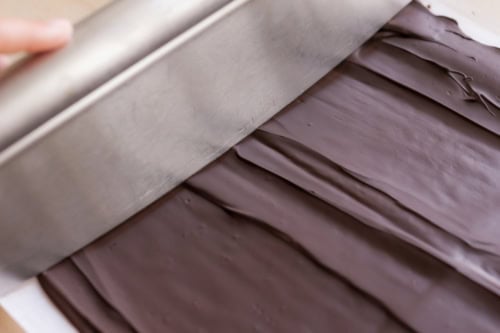 A large metal scraper smooths out a layer of dark chocolate on a flat surface.