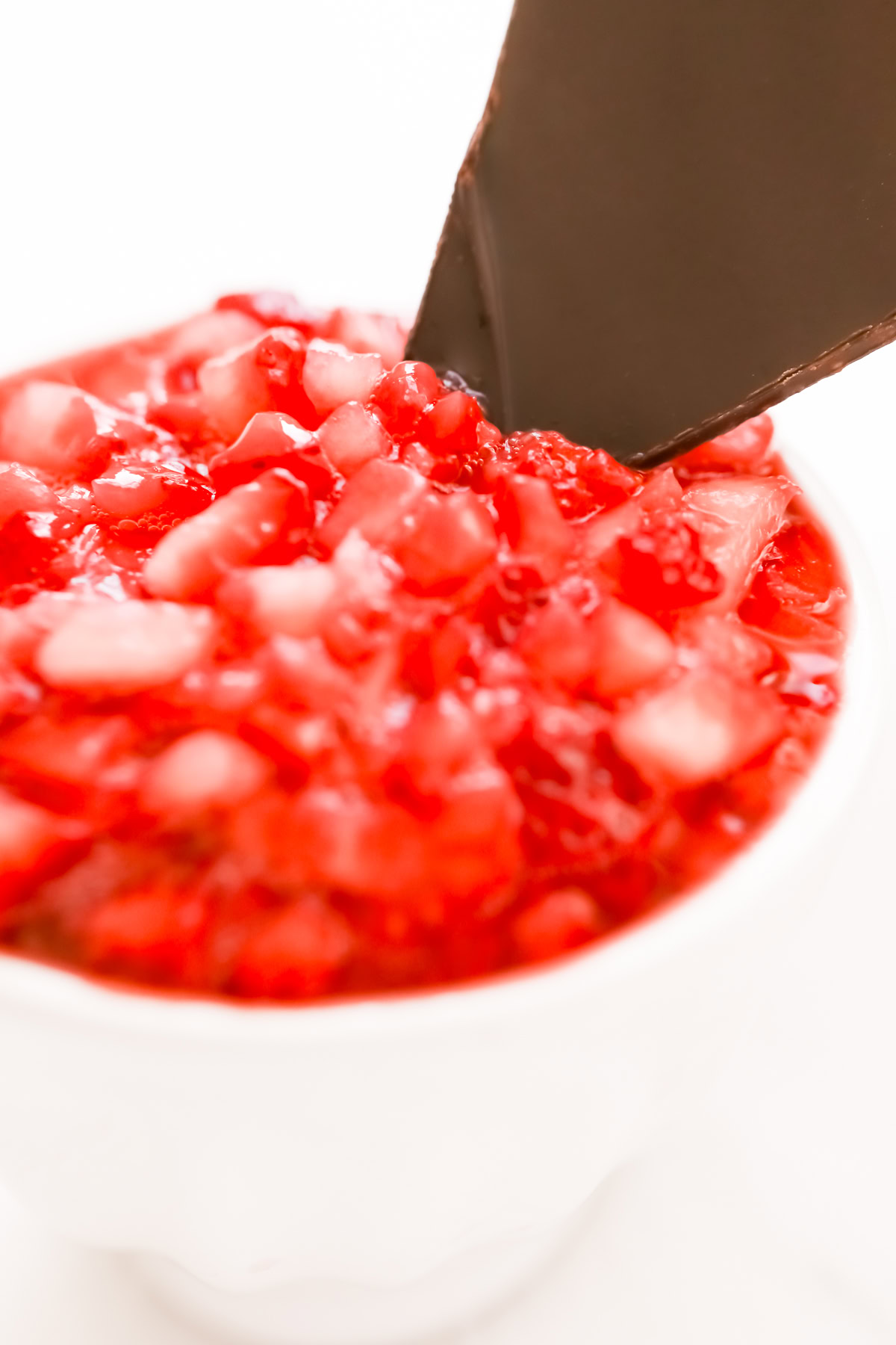A close-up of diced strawberries mixed with small ice cubes in a white cup. A piece of dark chocolate is inserted into the mixture.