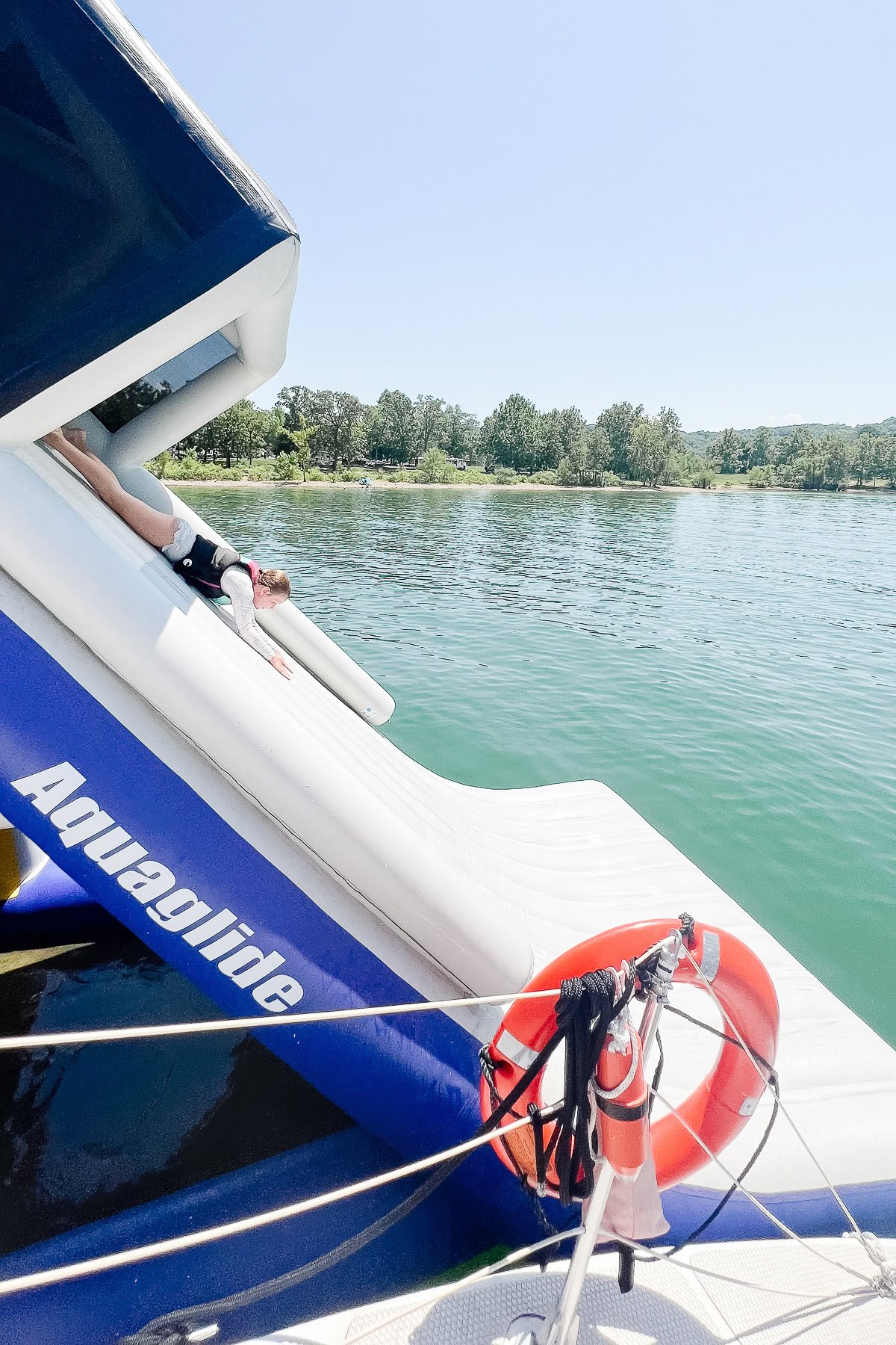 A person slides down an inflatable water slide labeled "Aquaglide" into a lake, with trees and greenery on the far shore—a fun example of things to do in Branson.