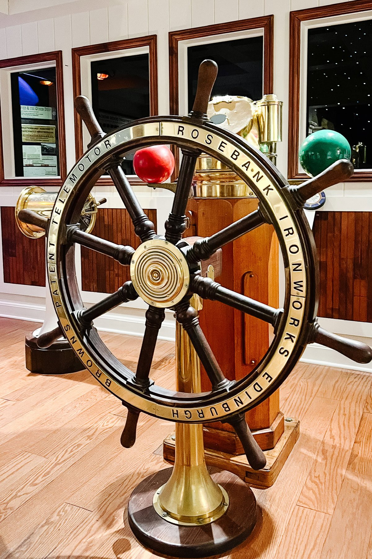 A large brass and wood ship’s wheel labeled "Rosebank Ironworks Edinburgh" is displayed indoors on a wooden floor, offering nautical decor for visitors seeking unique things to do in Branson. Mirrors in the background enhance its maritime charm.