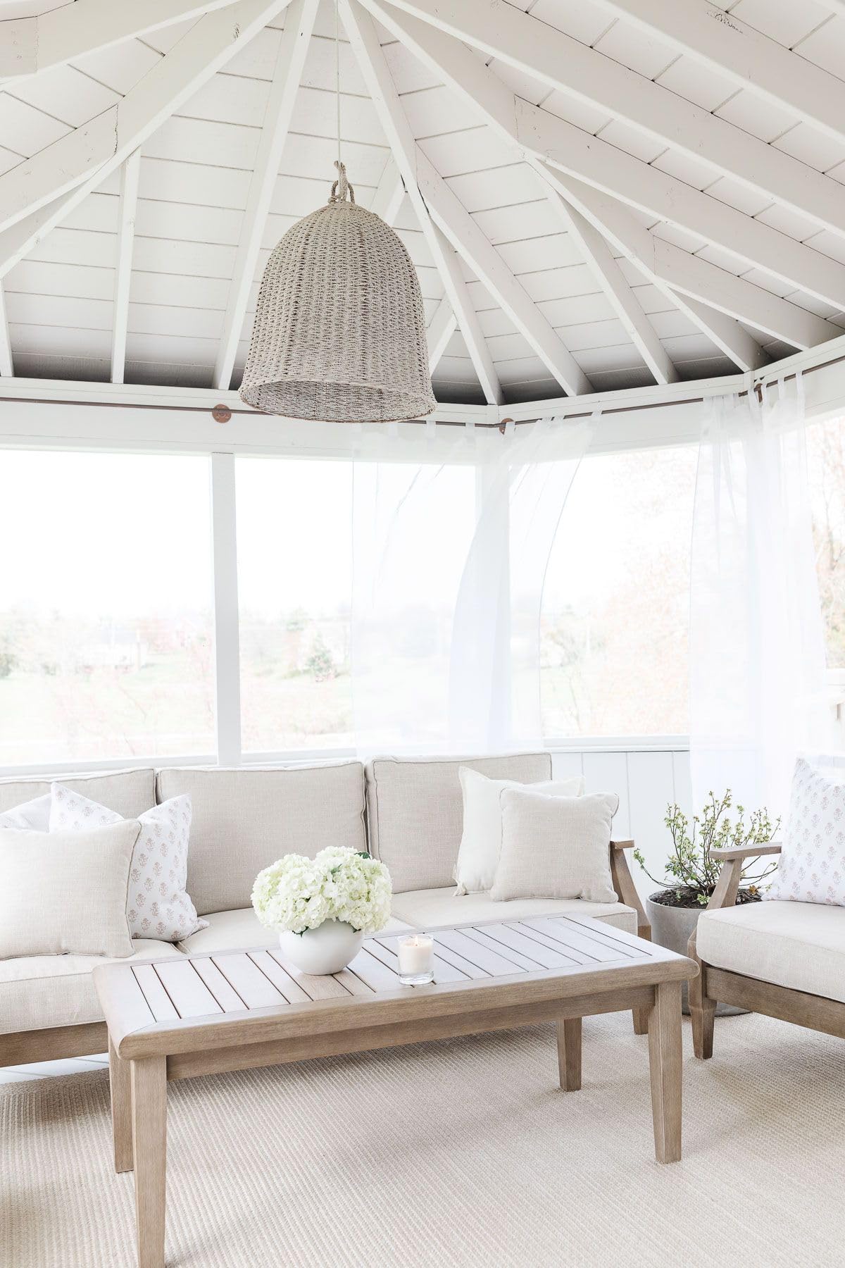 A bright, airy sunroom with white walls and ceiling, light wood furniture, neutral cushions, a woven pendant light, and a coffee table with flowers and a candle—perfect for relaxing or shopping Prime Day deals.
