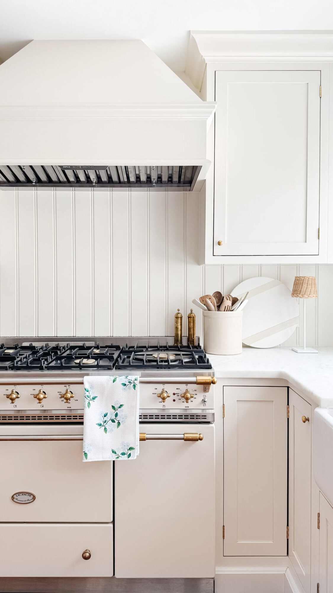 A cream-colored kitchen with a gas stove, hood vent, white cabinetry, and gold accents—perfect for cooking up Prime Day deals—with a towel featuring a green floral pattern hanging on the oven door.