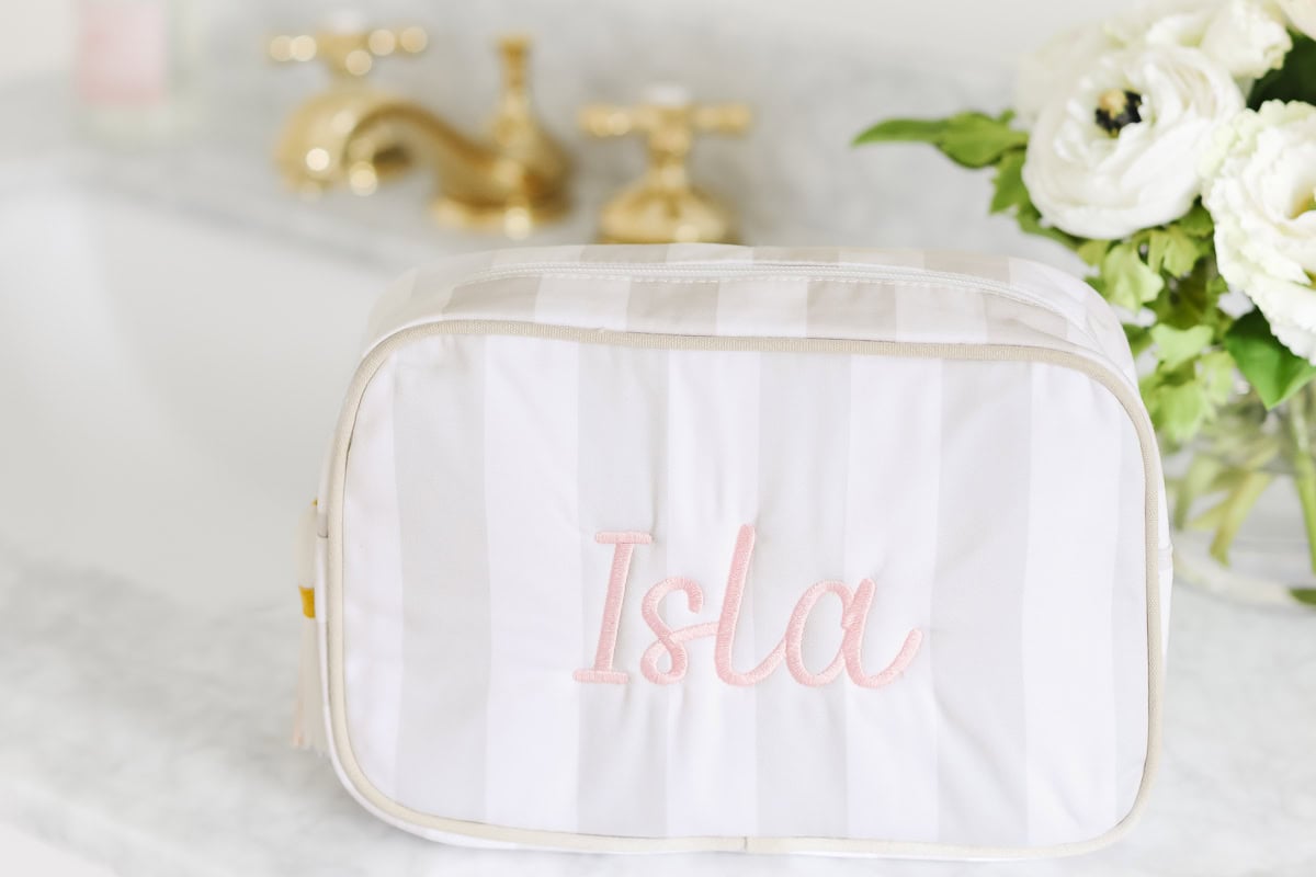 A striped white toiletry bag with the name "Isla" embroidered in pink sits on a marble countertop near a sink and white flowers, making it a chic pick for Prime Day.